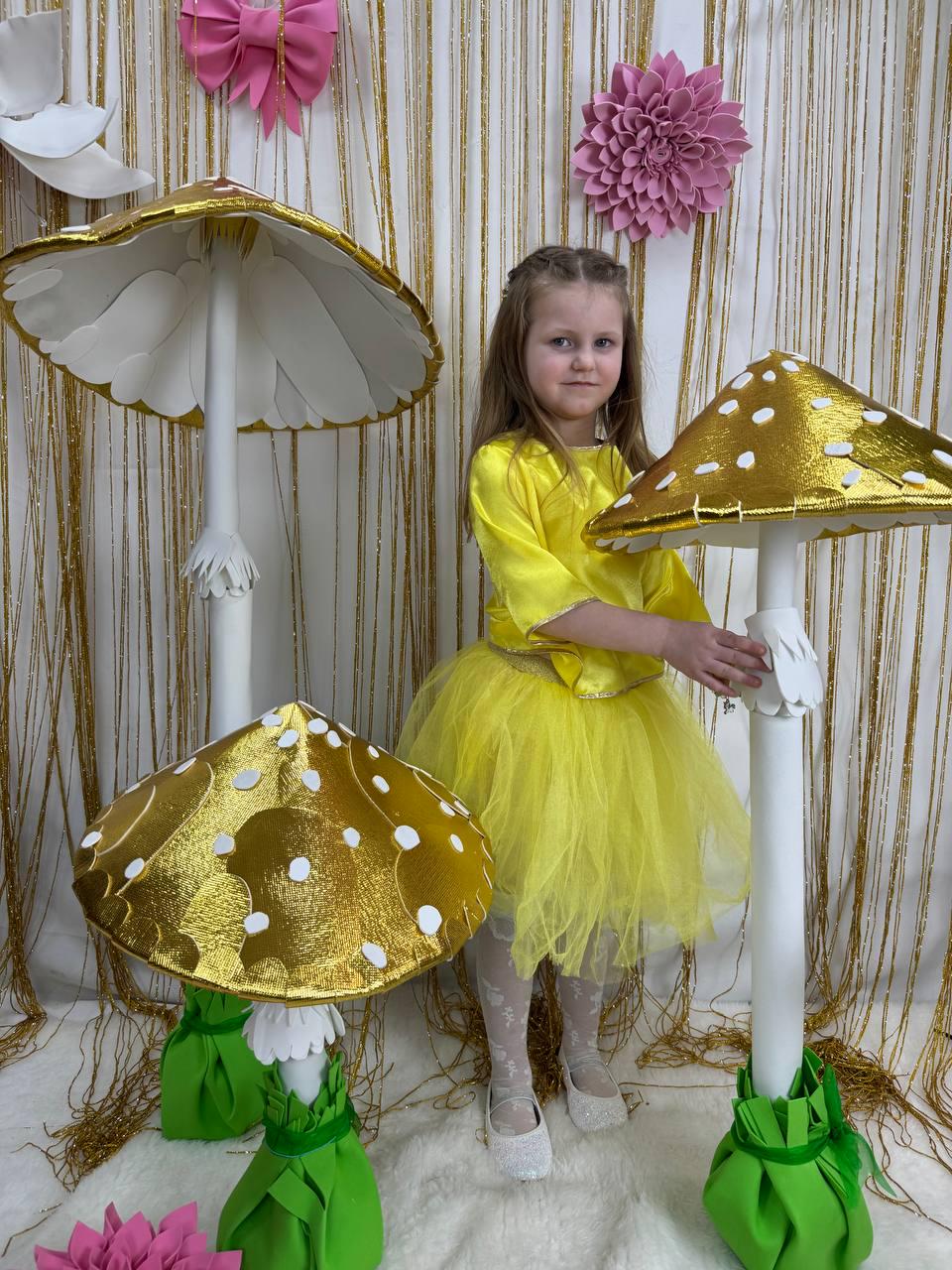 May include: Three large, white and gold mushroom decorations with green bases. The mushrooms are made of foam and have a shiny, metallic finish. A young girl in a yellow dress is standing in front of the mushrooms.