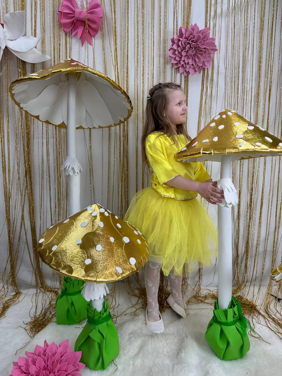May include: Three large golden mushroom decorations with white stems and green bases. The mushrooms are arranged in a row, with the largest mushroom in the back and the smallest mushroom in the front. A young girl in a yellow dress is standing next to the mushrooms.