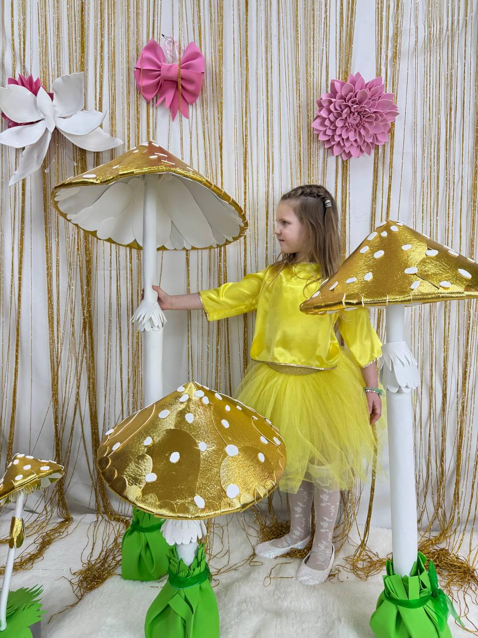 May include: A young girl in a yellow dress stands in front of a backdrop of gold fringe and pink flowers. She is reaching out to touch a large white mushroom with a gold top. There are smaller gold mushrooms with white dots on the tops in the foreground.