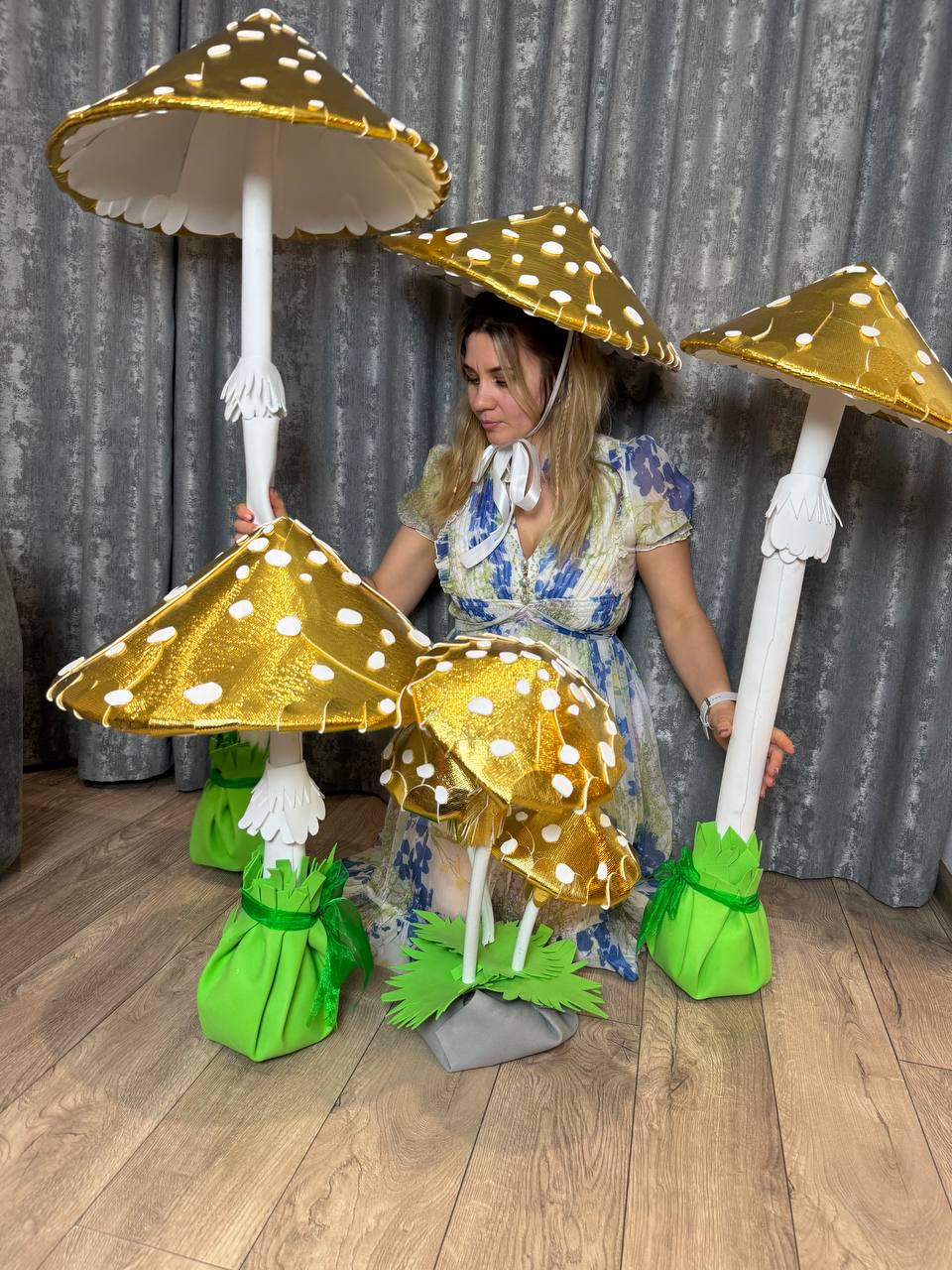 May include: Four large golden mushroom decorations with white polka dots. The mushrooms have white stems and green bases. A person is wearing a golden mushroom hat with white polka dots.