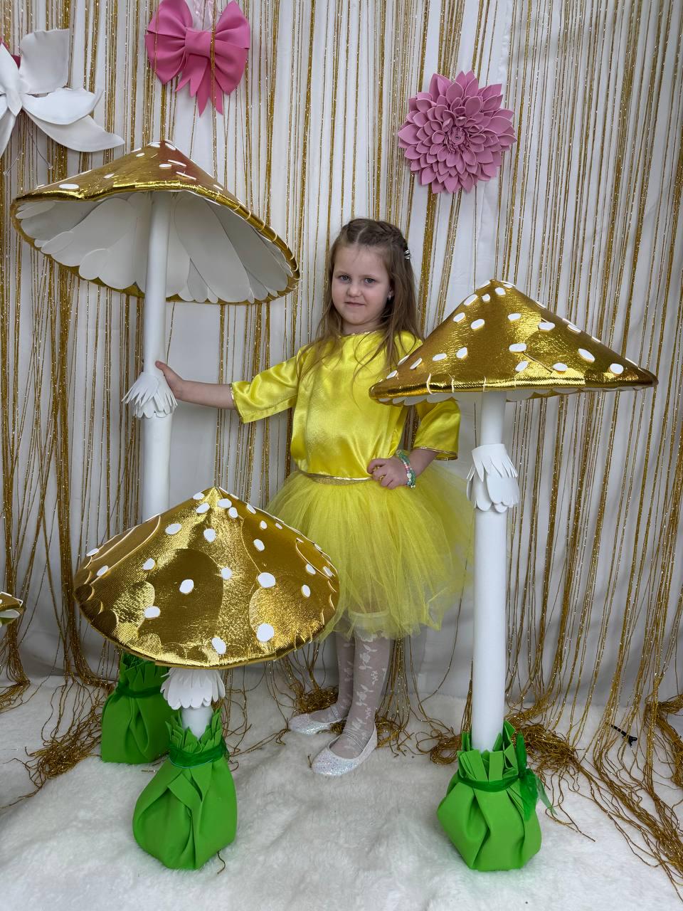 May include: Three large, decorative, golden mushroom props with white polka dots. The mushrooms have green bases and white stems. A young girl is standing next to the largest mushroom.