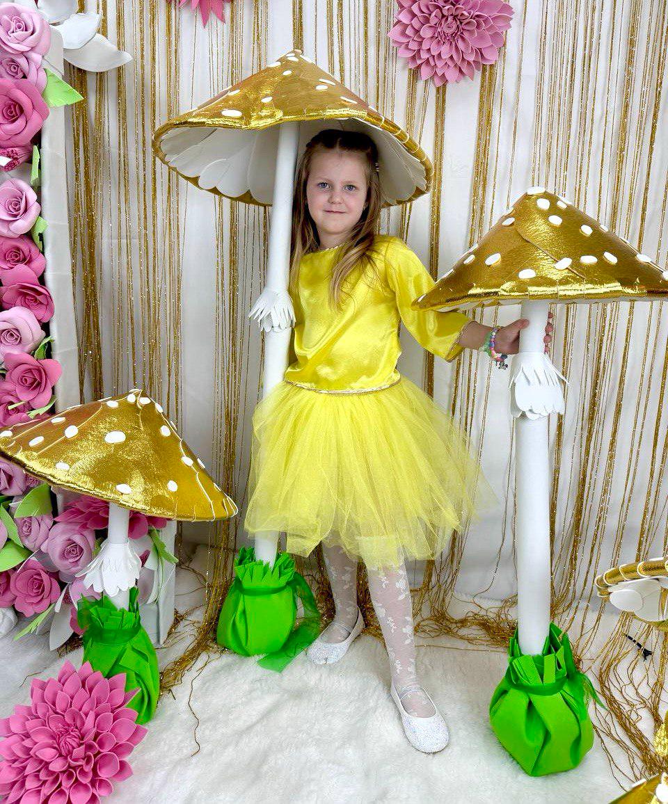 May include: A young girl wearing a yellow dress and white tights stands in front of a backdrop of gold and white streamers. She is holding a large, gold and white mushroom prop. There are two other mushroom props in the background.