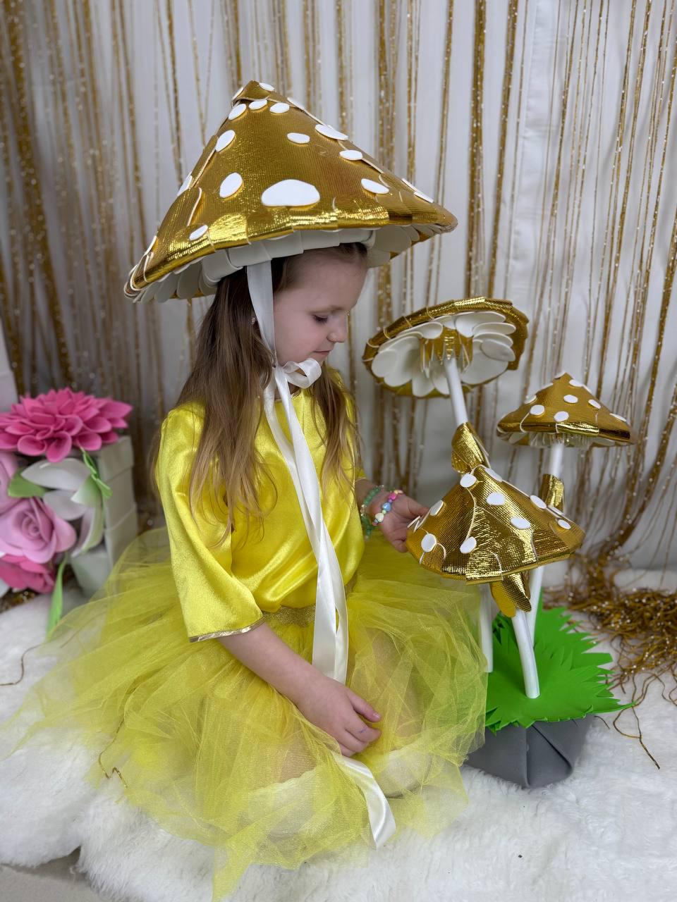 May include: A young girl wearing a yellow tulle skirt and a gold mushroom hat with white polka dots. She is holding a large gold mushroom prop with white polka dots.