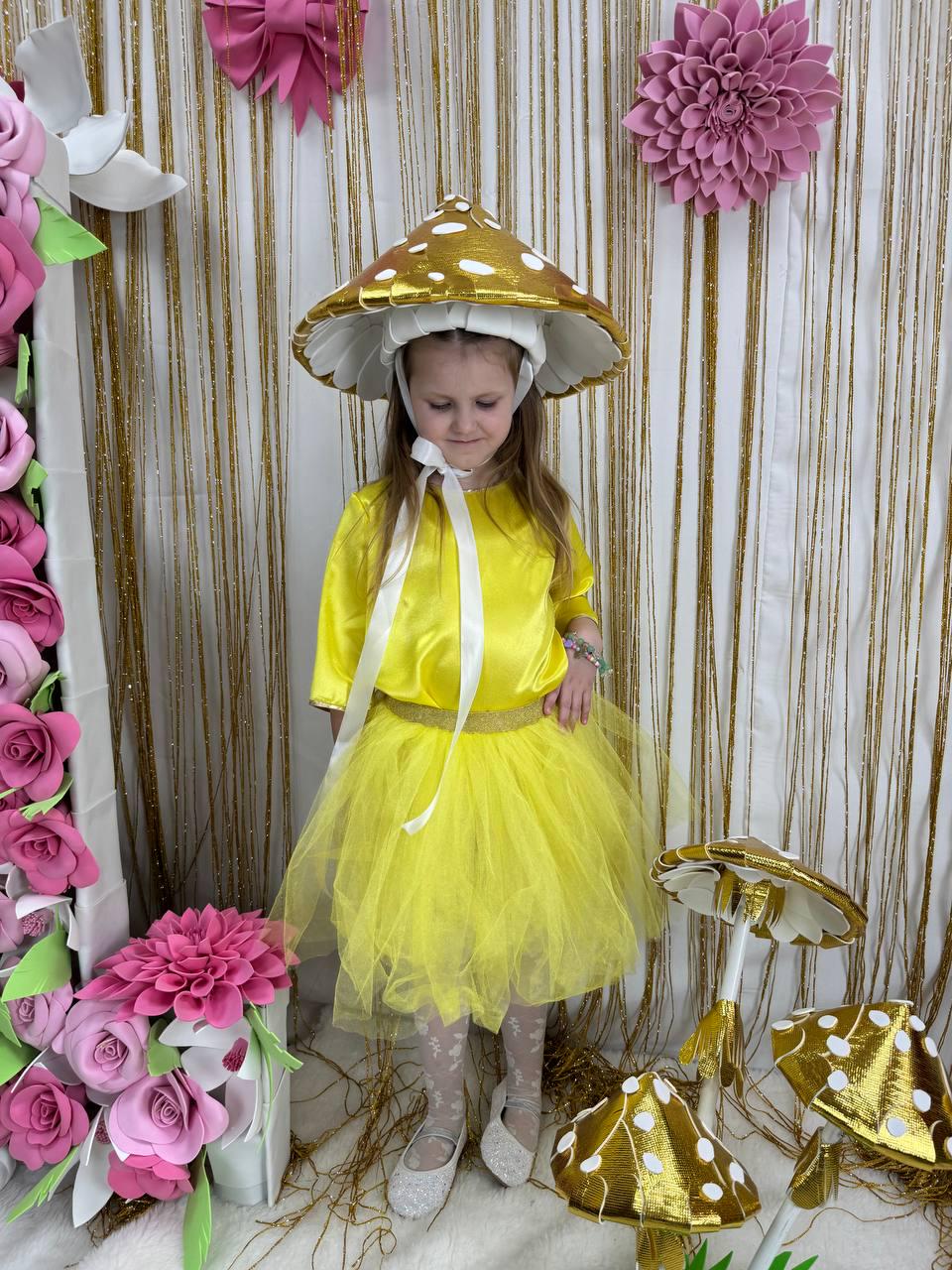 May include: A young girl wearing a yellow dress and a white mushroom hat with gold accents. She is standing in front of a backdrop of pink flowers and gold streamers. There are also three gold mushroom decorations on the floor.