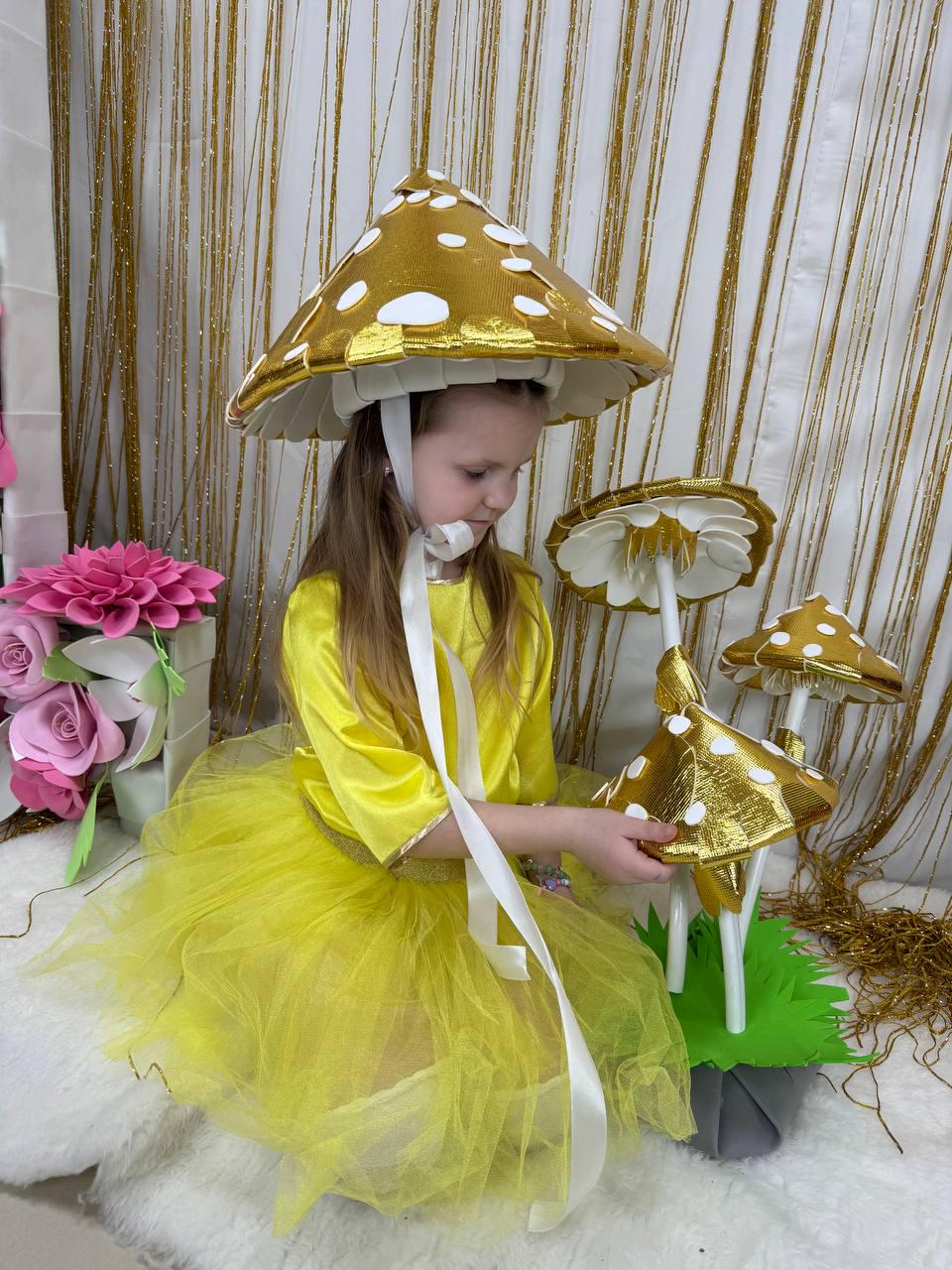 May include: A young girl wearing a yellow dress and a gold and white mushroom hat. She is holding a large gold and white mushroom prop.