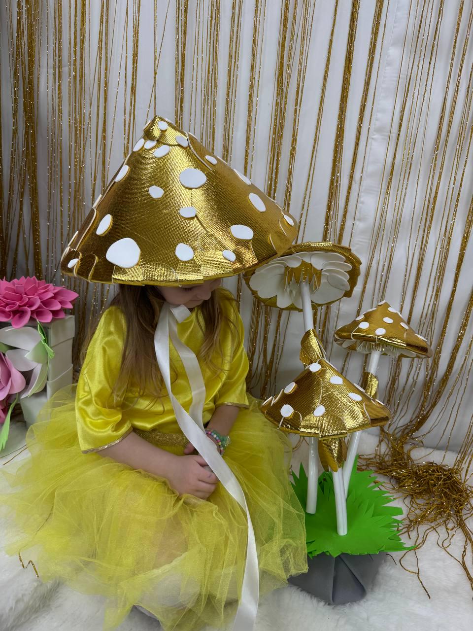 May include: A child wearing a yellow dress and a gold mushroom hat sits in front of a set of three gold and white mushroom decorations. The mushrooms are on a green base.