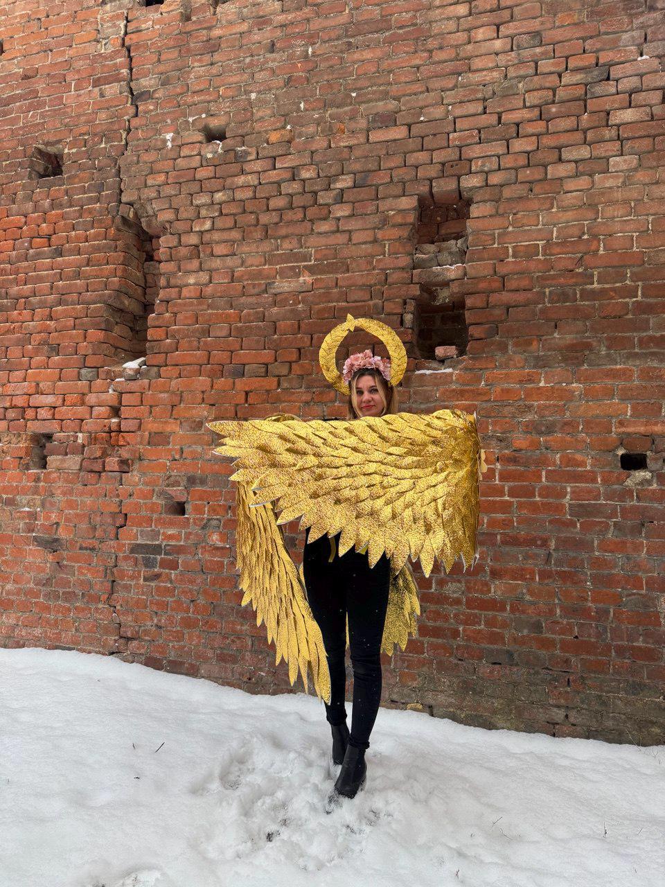 May include: A person wearing a gold glitter angel wing costume with a flower crown. The person is standing in front of a brick wall with snow on the ground.