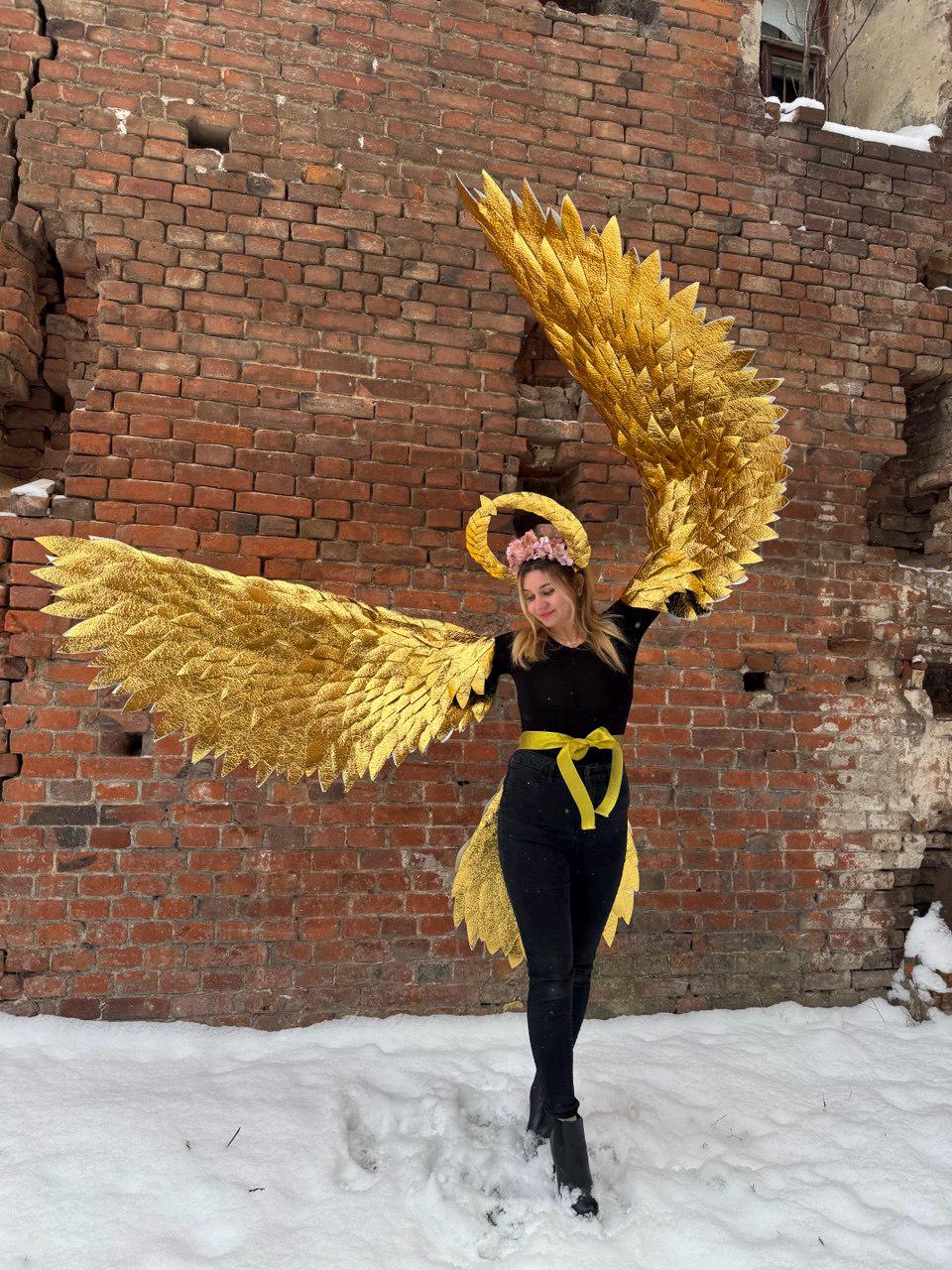 May include: A person wearing a black outfit with a yellow sash and a gold halo, holding large gold wings in front of a brick wall.