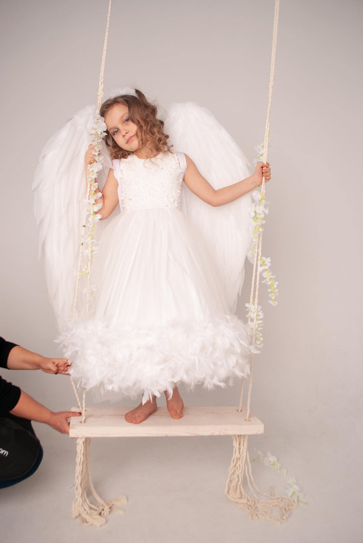 May include: A young girl dressed as an angel wearing a white dress with feather trim, sits on a white wooden swing with white rope. The girl is holding onto the rope with one hand and has white angel wings.