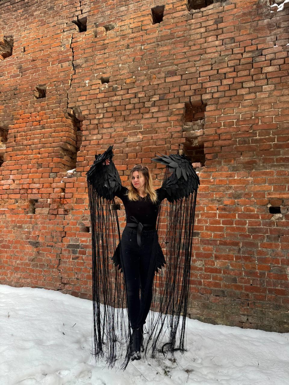 May include: A person wearing a black outfit with long, black, fringed wings. The wings are made of fabric and have a feathered appearance. The person is standing in front of a brick wall.