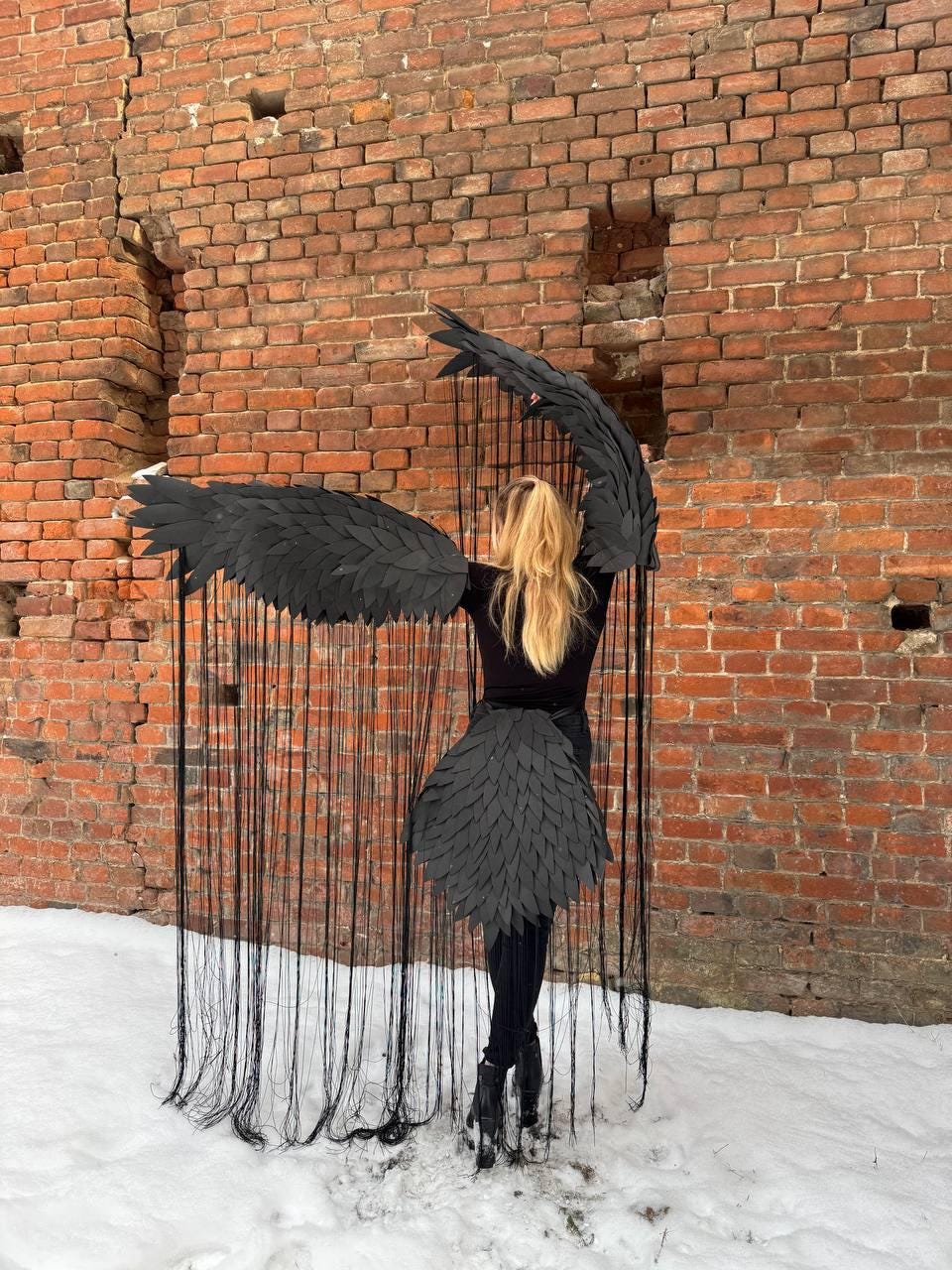 May include: A person wearing a black costume with large black wings made of feathers. The wings are attached to a black skirt and the person is standing in front of a brick wall. The background is covered in snow.