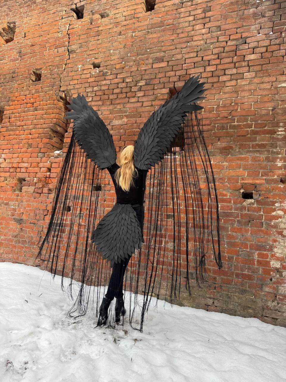 May include: A person wearing a black costume with large black wings and a long black skirt. The wings are made of feathers and the skirt is made of fabric. The person is standing in front of a brick wall.