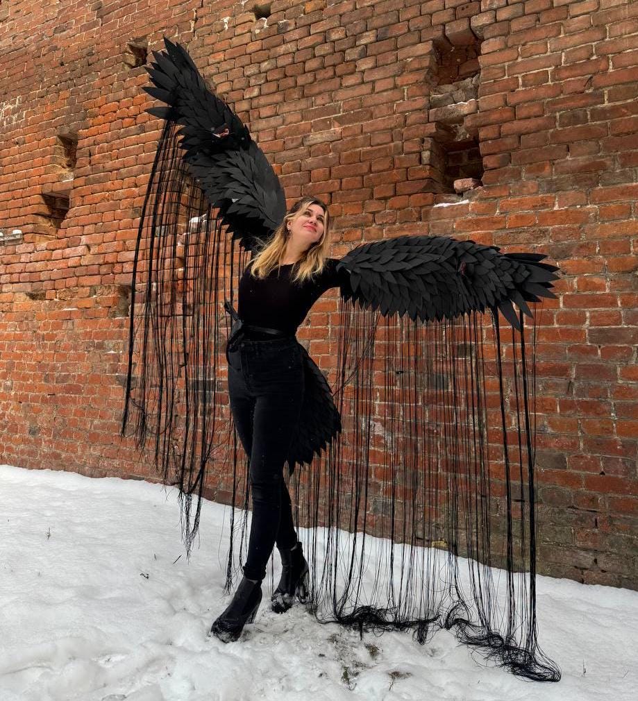 May include: A person wearing a black outfit with large black wings made of feathers and fringe. The wings are spread out behind the person, and the person is standing in front of a brick wall. The person is standing in the snow.