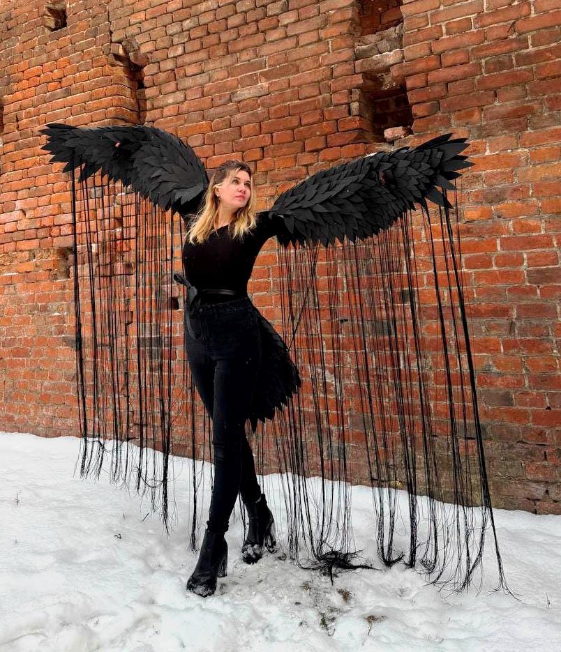 May include: A person wearing a black outfit with long black wings made of feathers and fringe. The wings are spread out behind the person, and the fringe is flowing in the wind. The person is standing in front of a brick wall.