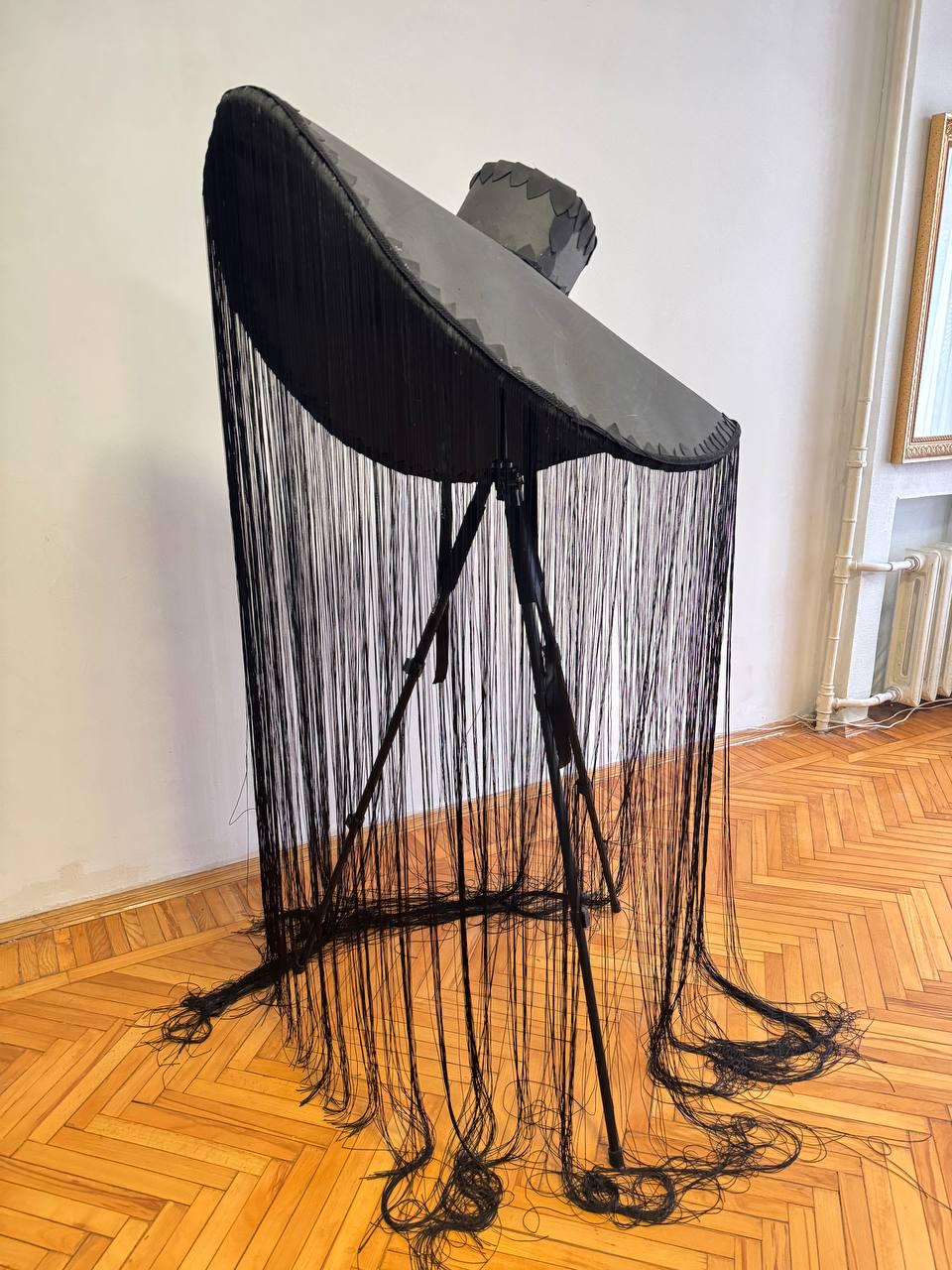 May include: A large black hat with a wide brim and long fringe hangs from a tripod stand. The hat is made of a dark fabric and has a slightly textured surface. The fringe is made of thin black threads that hang down to the floor.