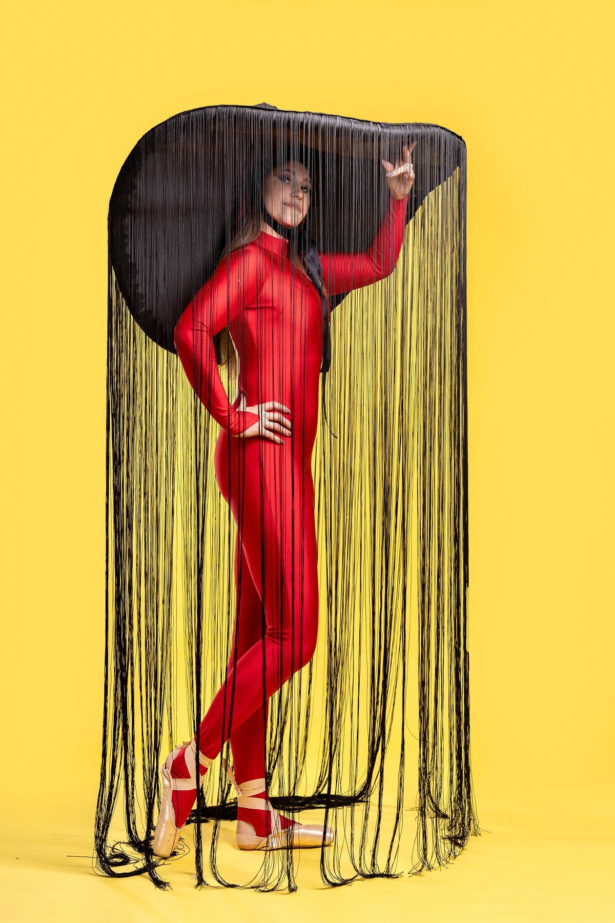 May include: A person wearing a red bodysuit and a large black hat with long black fringe. The hat is covering the person's head and shoulders. The person is standing on a yellow background.