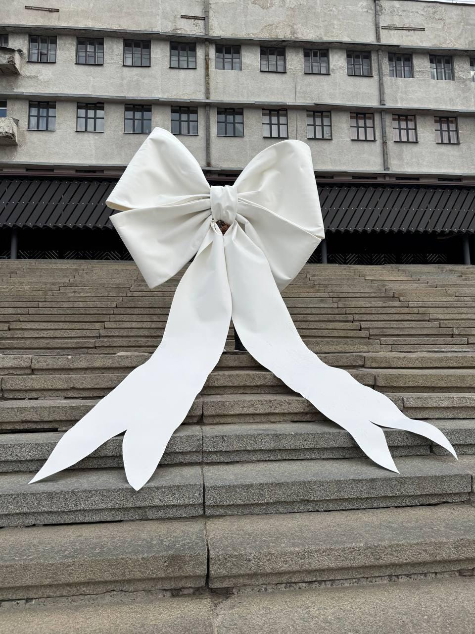 May include: A large white bow is placed on a set of concrete stairs in front of a building. The bow is made of fabric and has a large, rounded center.
