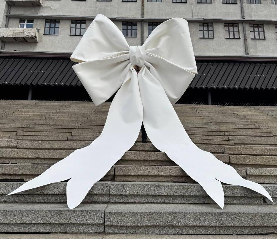 May include: A large white bow made of fabric is placed on a set of concrete steps. The bow is tied in a knot and the tails of the bow are long and flowing.