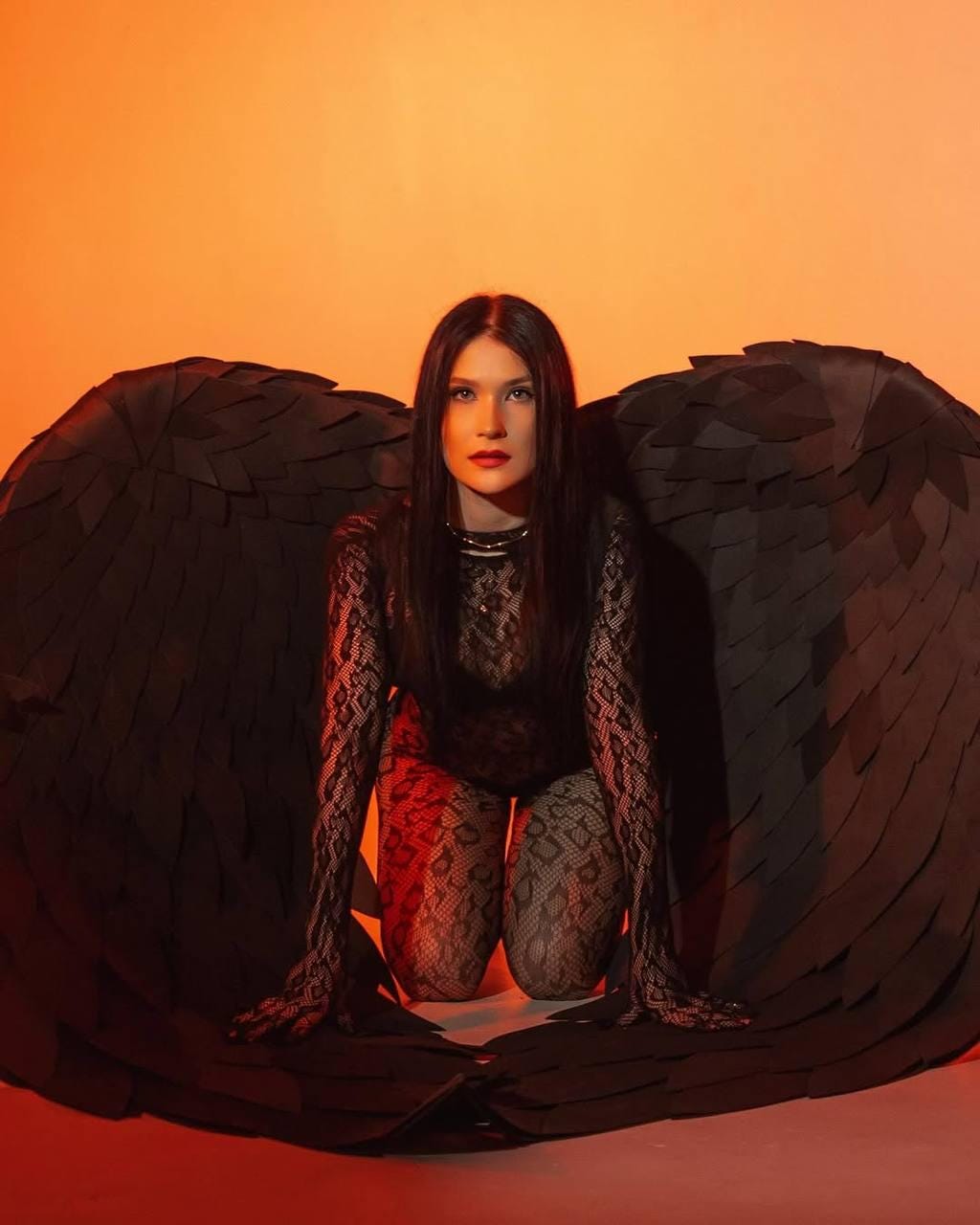 May include: A woman wearing a black lace bodysuit and fishnet stockings kneels in front of a pair of large black angel wings.