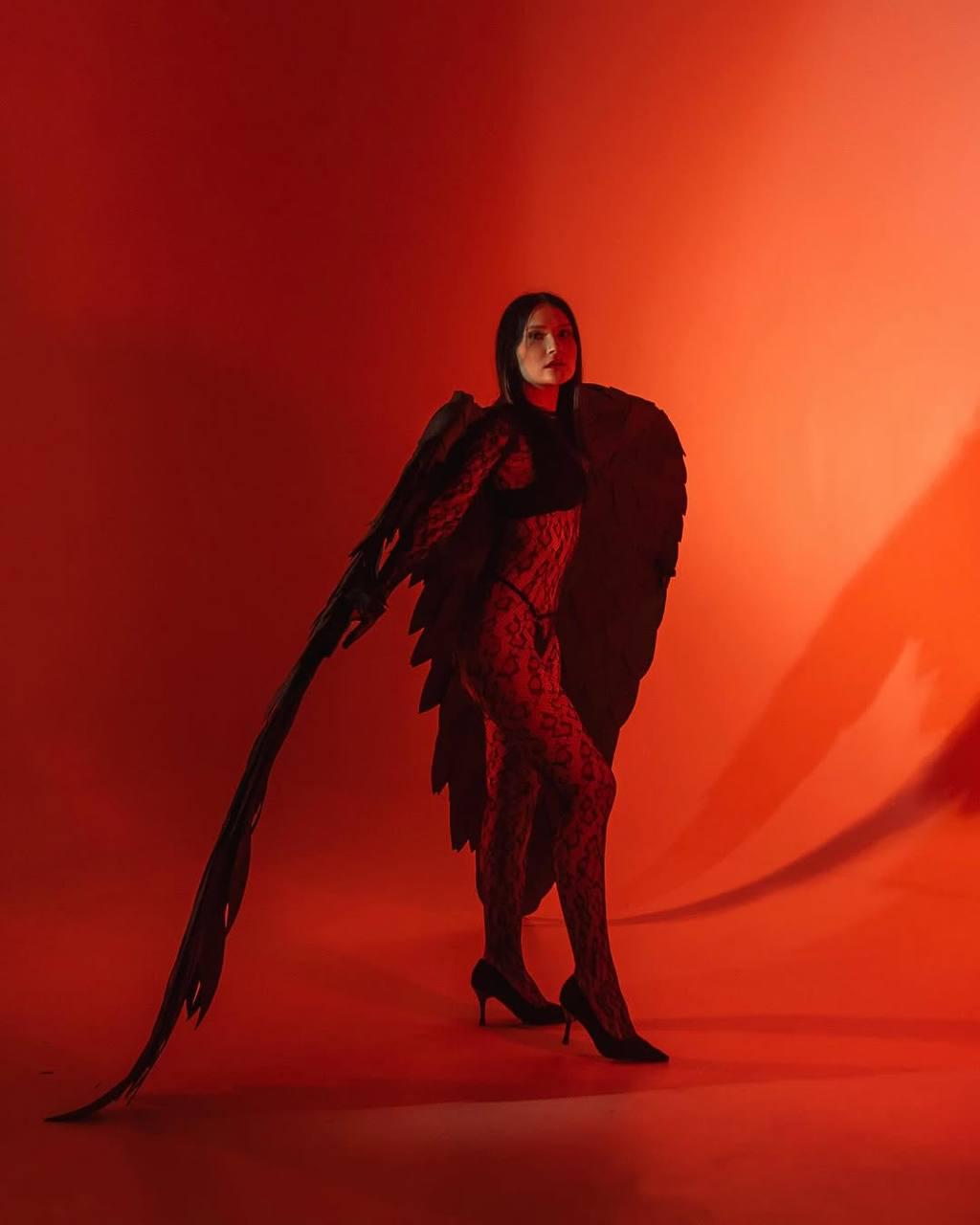May include: A woman wearing a black lace bodysuit with black wings and black heels stands against a red background.
