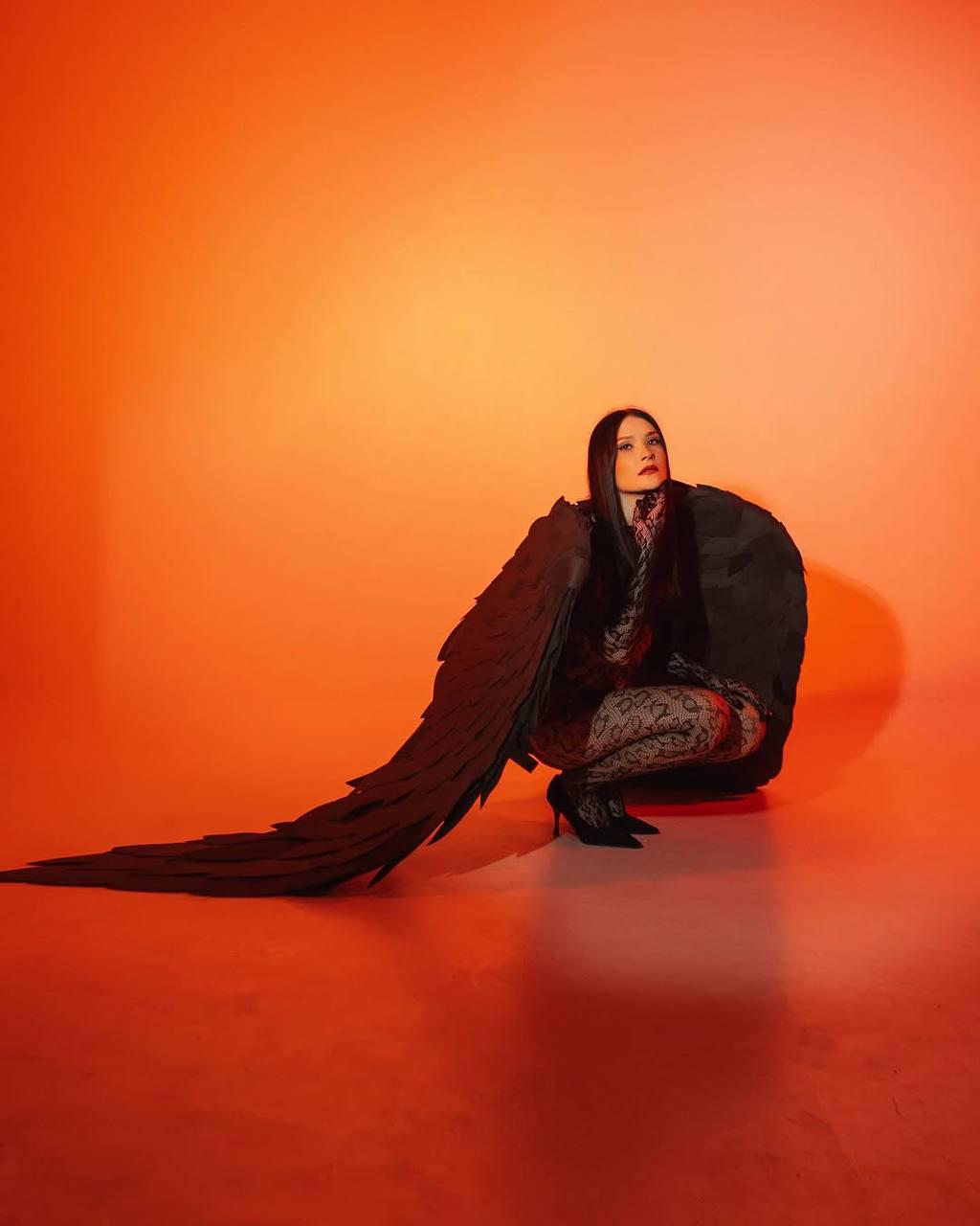 May include: A woman wearing a black outfit with large black wings sits on an orange background. The wings are made of feathers and are spread out behind her.