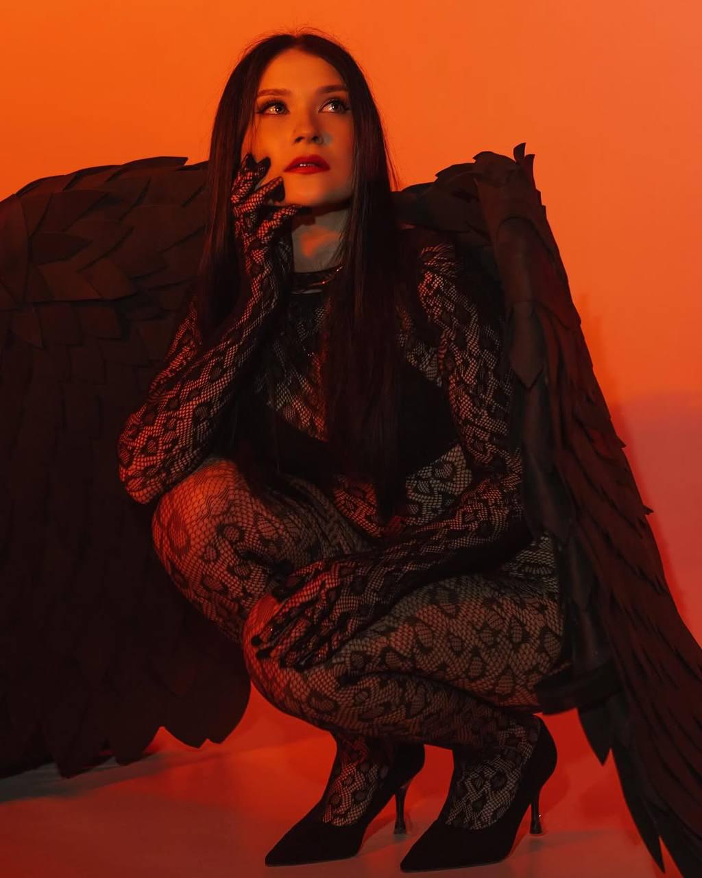 May include: A woman wearing a black lace bodysuit and gloves poses with large black angel wings against a red background.