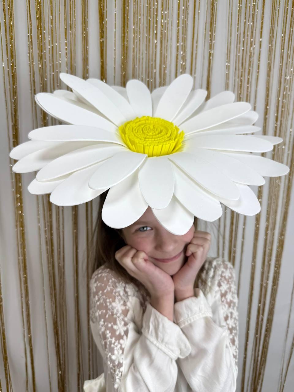 May include: A large white paper daisy flower with a yellow center. The flower is being worn as a hat by a child.