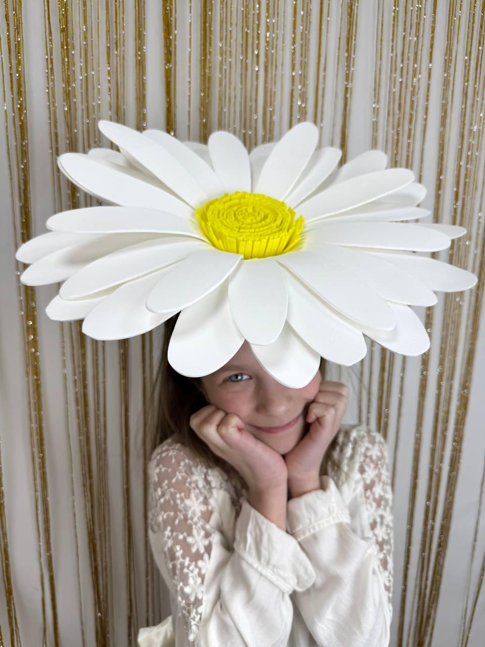 May include: A large, white daisy flower hat with a yellow center. The hat is worn by a person, with the petals extending outwards. The background features a gold, glittery curtain.