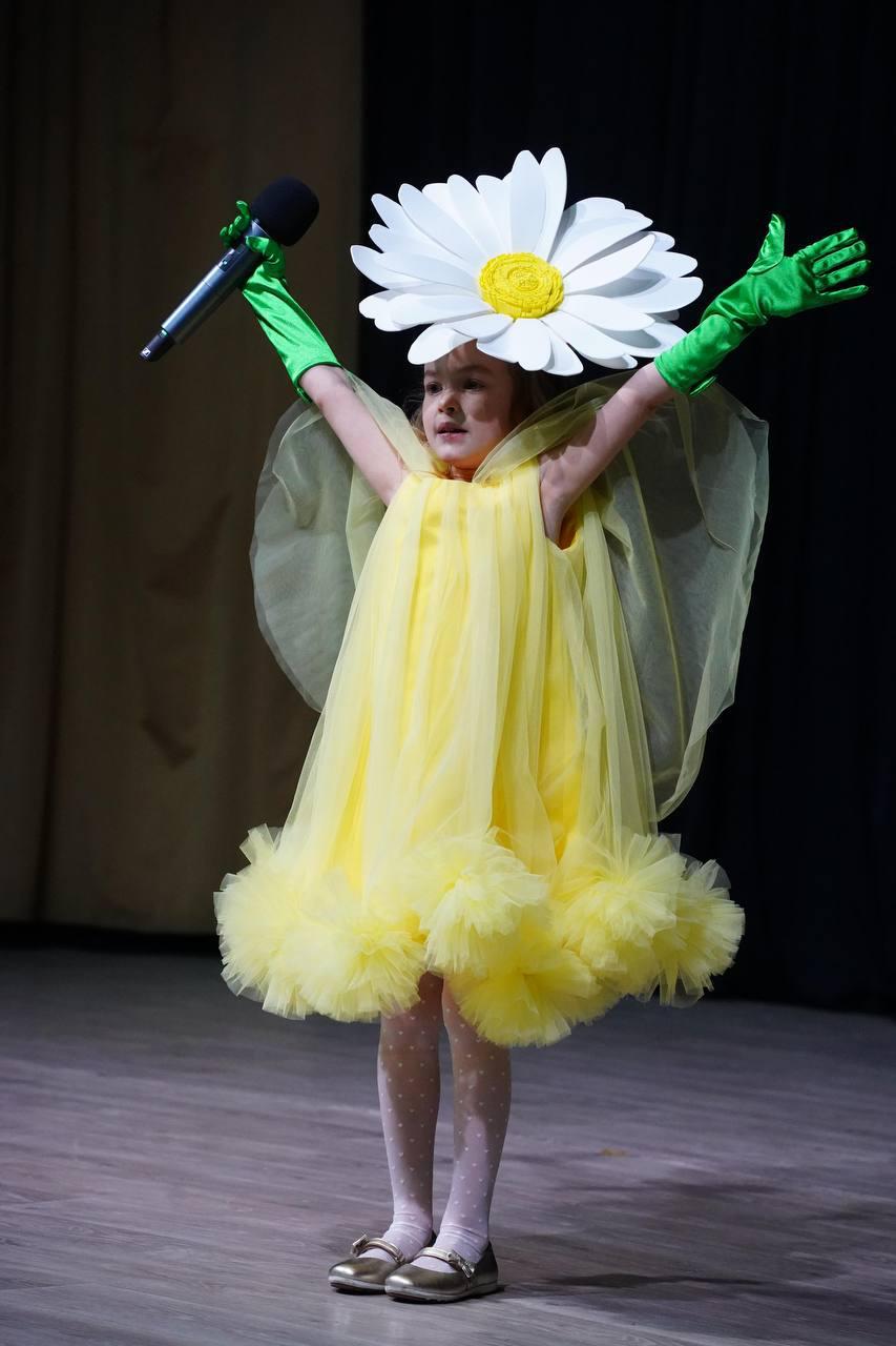 May include: A young girl wearing a yellow tulle dress with a large white daisy flower on her head. She is holding a microphone and has her arms raised in the air.