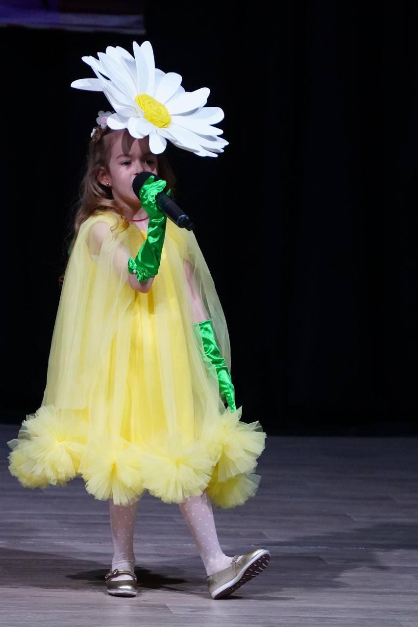 May include: A young person in a yellow dress with a large white daisy headpiece and green gloves, holding a microphone. The dress has a ruffled hem and a sheer cape. The background is dark.