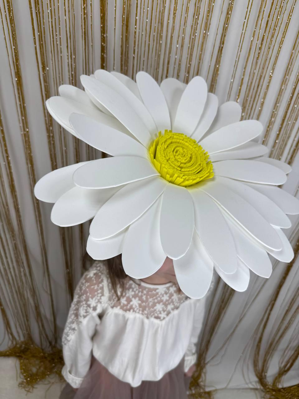 May include: A large, white daisy flower headpiece with a bright yellow center. The oversized floral accessory has multiple layers of white petals. The headpiece is worn by a person wearing a white lace top and a pink skirt.