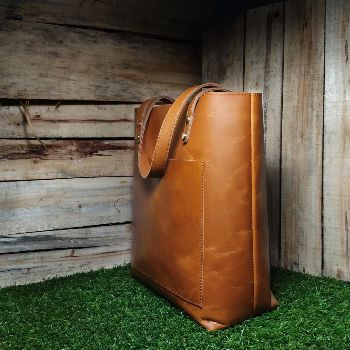 May include: A brown leather tote bag with a front pocket and two straps. The bag is sitting on a green grassy surface.