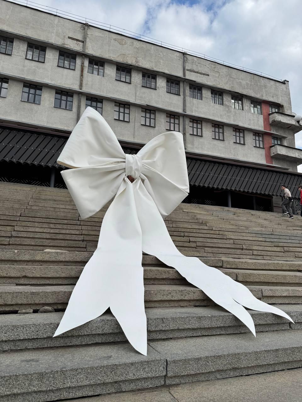 May include: A large white bow is placed on a set of concrete steps in front of a building. The bow is made of fabric and has two long tails.