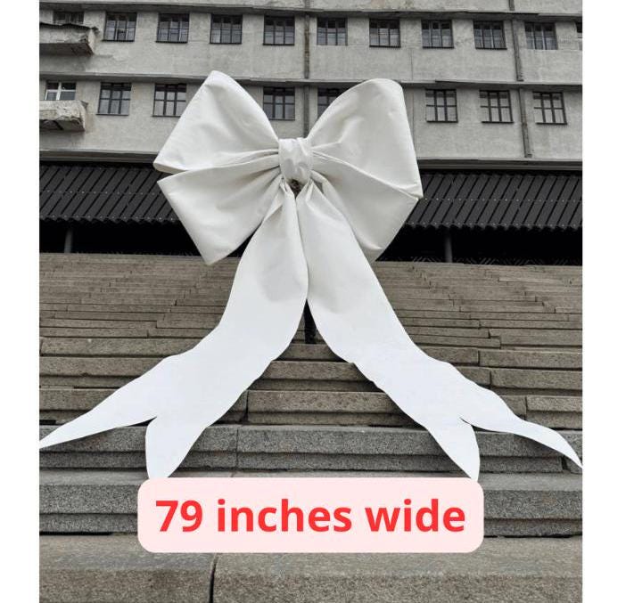 May include: A large white bow made of fabric, measuring 79 inches wide, is displayed on a set of concrete steps.
