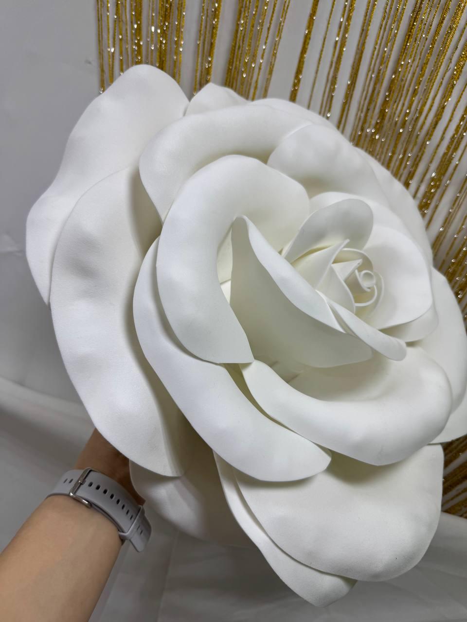 May include: A large, white, artificial rose with multiple layers of petals. The rose is held by a person. The background features gold glitter strands. This decorative flower is suitable for events and home decor.