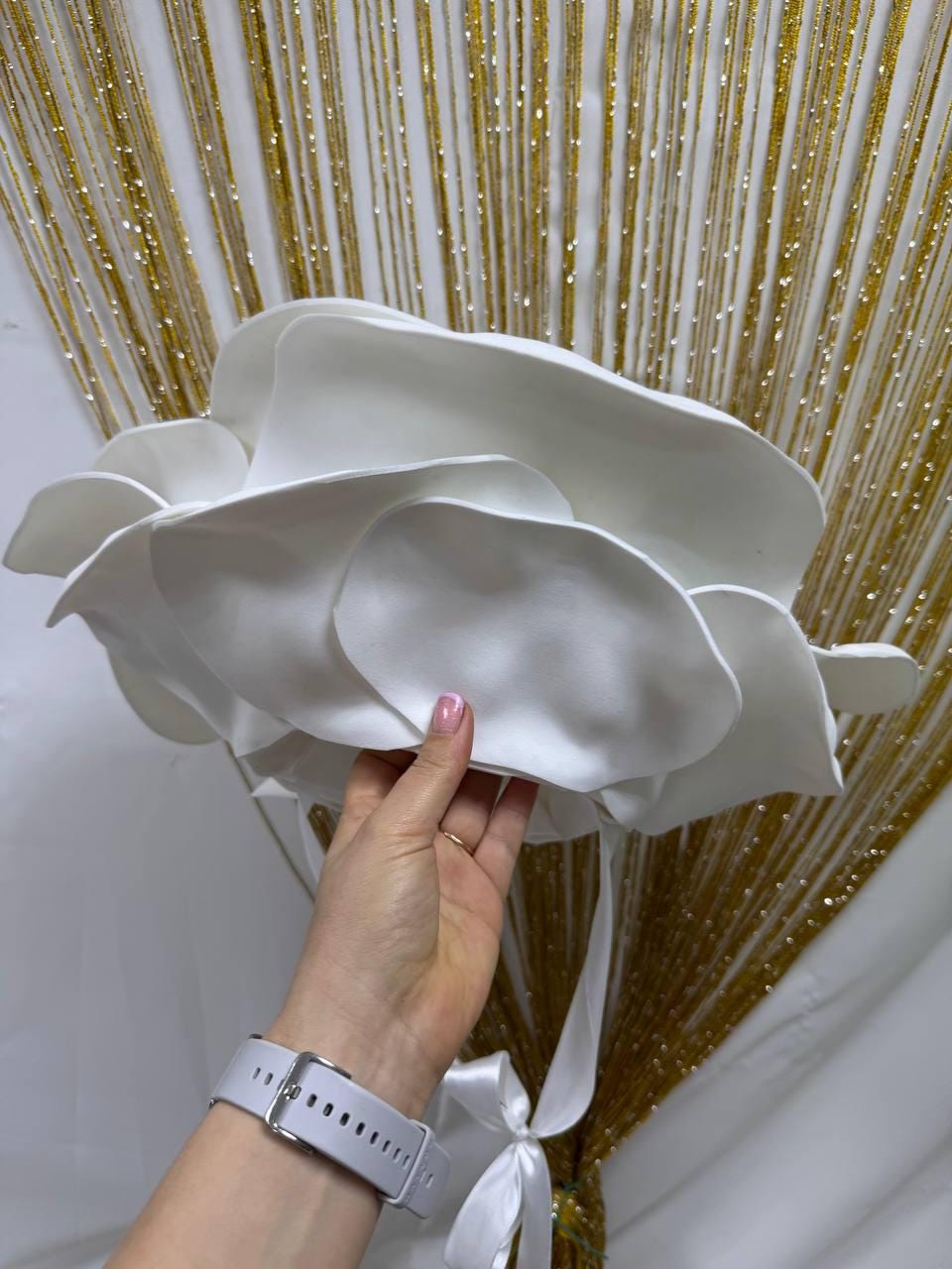 May include: A large, white, paper flower with layered petals. The flower is held by a hand, with white ribbons tied at the base. The background features gold strands against a white backdrop.