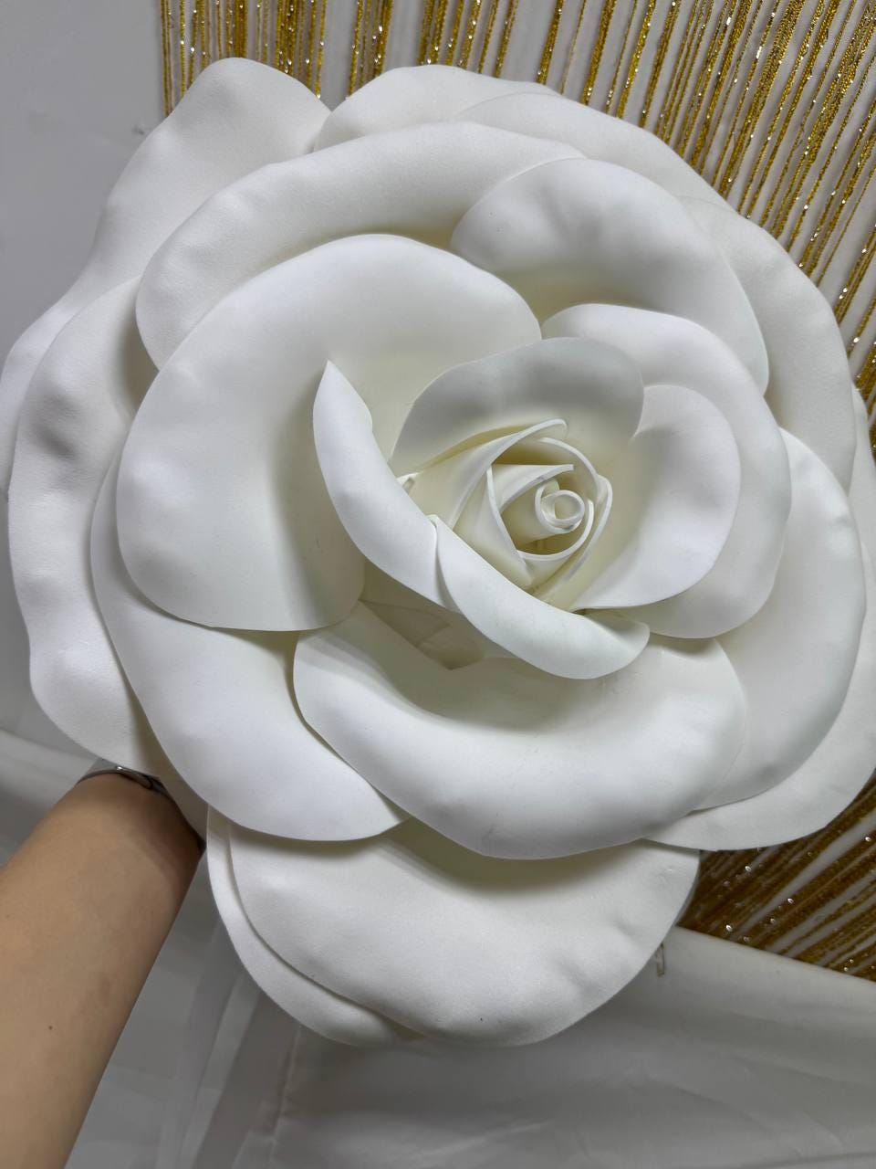 May include: A large, white, artificial rose with multiple layers of petals. The flower is in full bloom, with a detailed center and soft, rounded petals. The background features gold glitter accents.