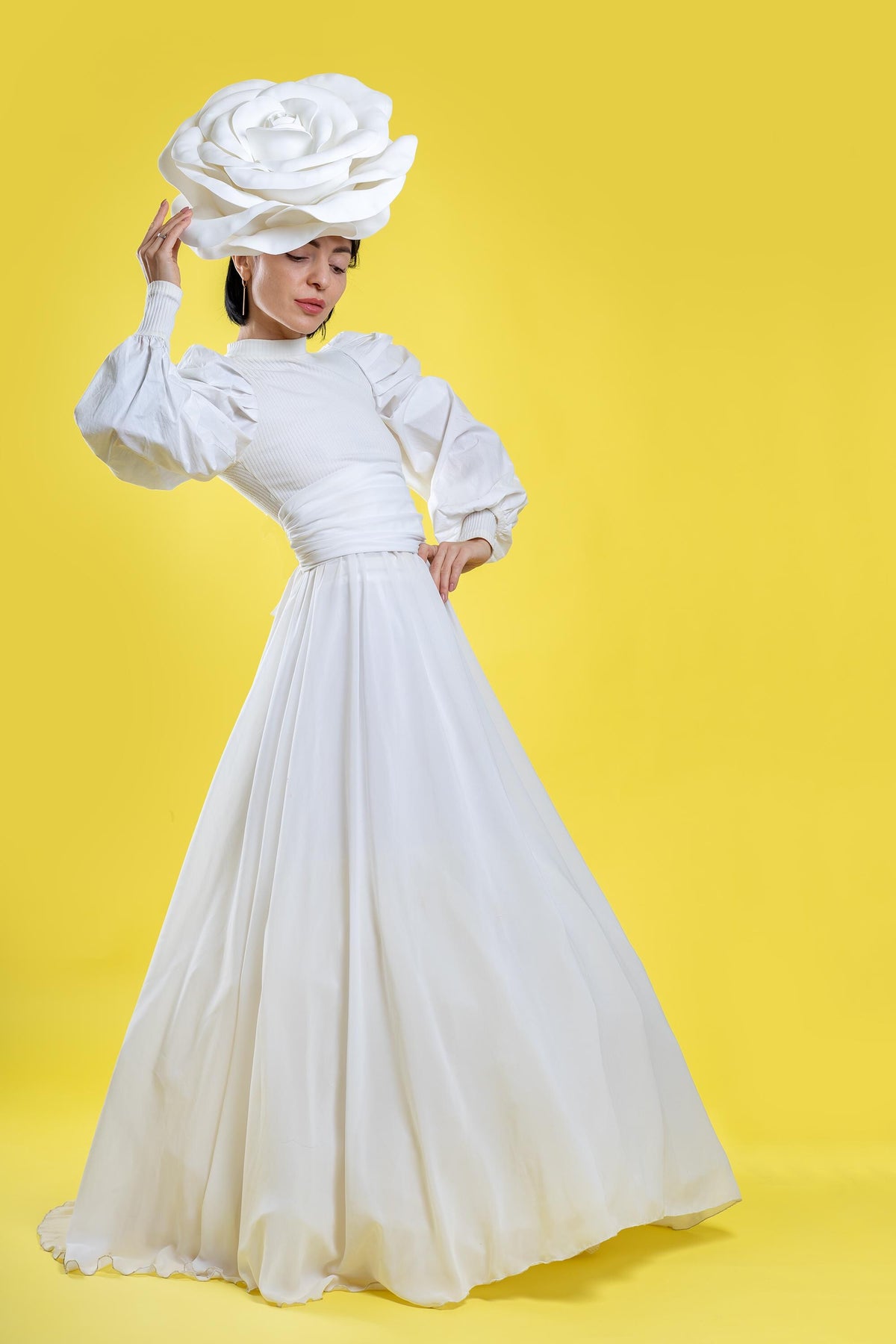 May include: A white dress with a high neckline and long, puffy sleeves. The dress features a cinched waist and a flowing skirt. A large, white rose-shaped headpiece is worn on the head. The background is a solid yellow.