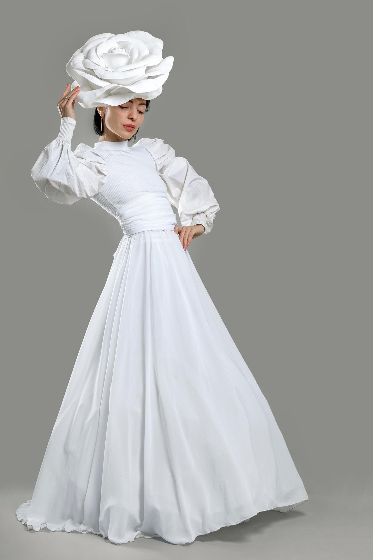 May include: A woman wearing a white dress and a large white rose-shaped hat. The dress has a high neckline, long sleeves with puffed shoulders, and a flowing skirt. The background is a solid gray.