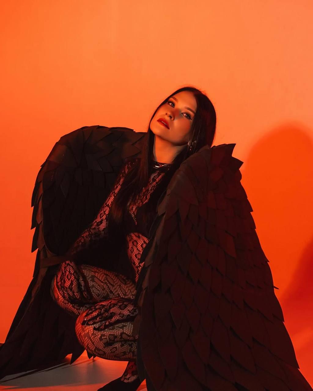 May include: A person wearing a black lace bodysuit and large black feathered wings, posing against a bright orange background.
