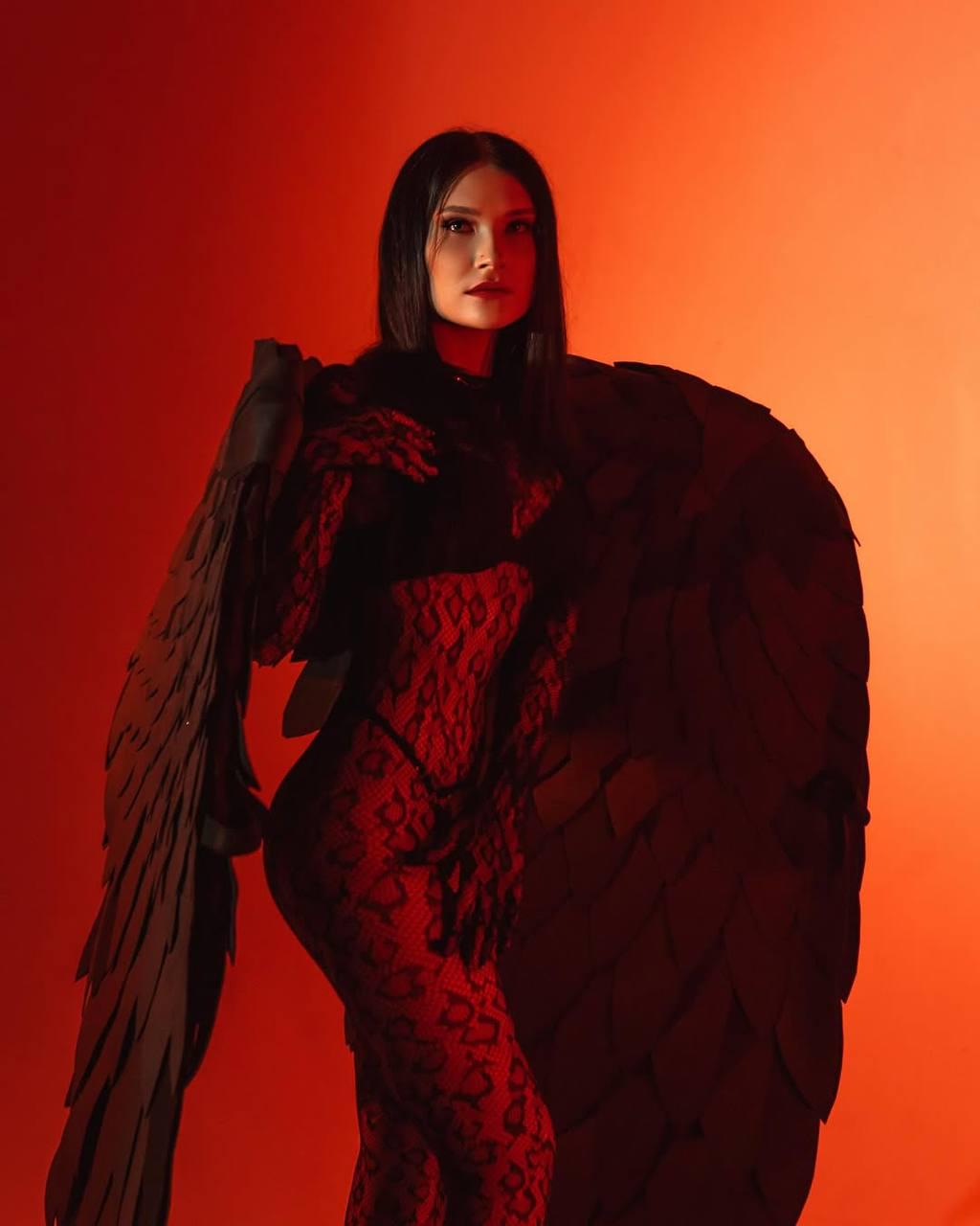 May include: A woman in a red lace bodysuit with black wings stands against a red background. The wings are made of a dark material and have a textured surface.