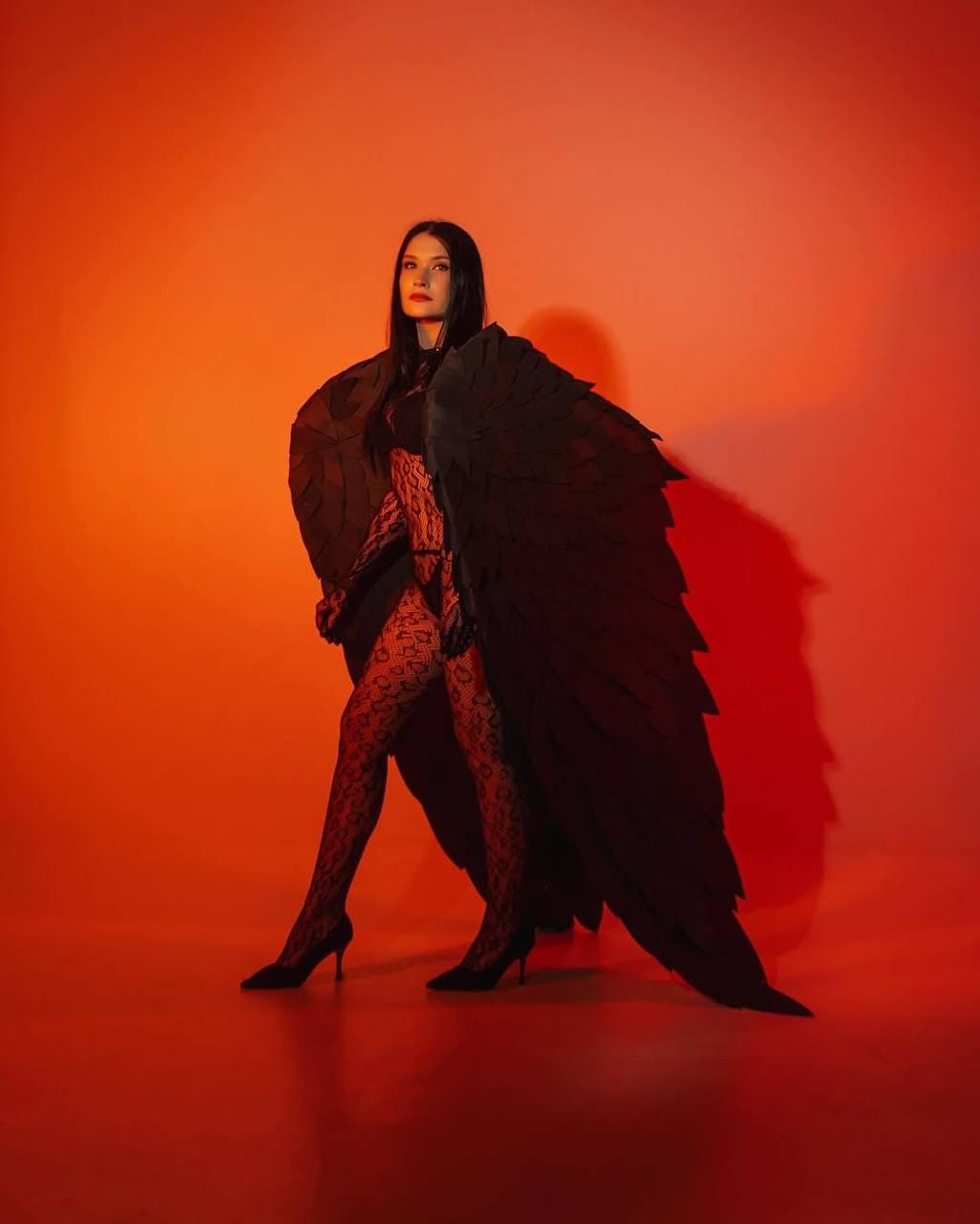 May include: A woman wearing a black lace bodysuit and black feathered wings poses in front of a red background.