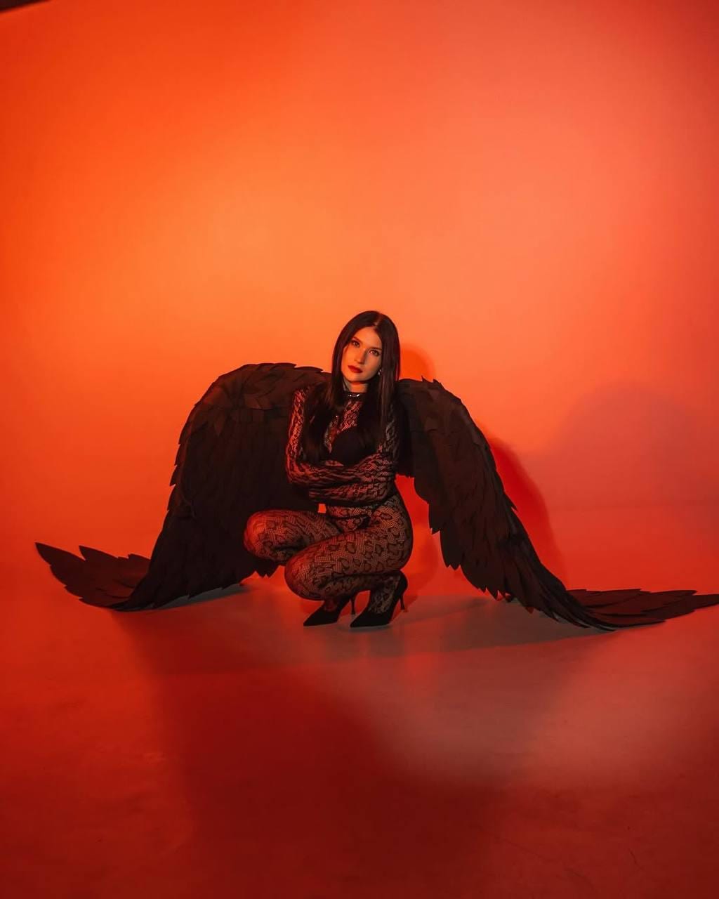 May include: A woman wearing a black lace bodysuit and large black angel wings poses in front of a red background.
