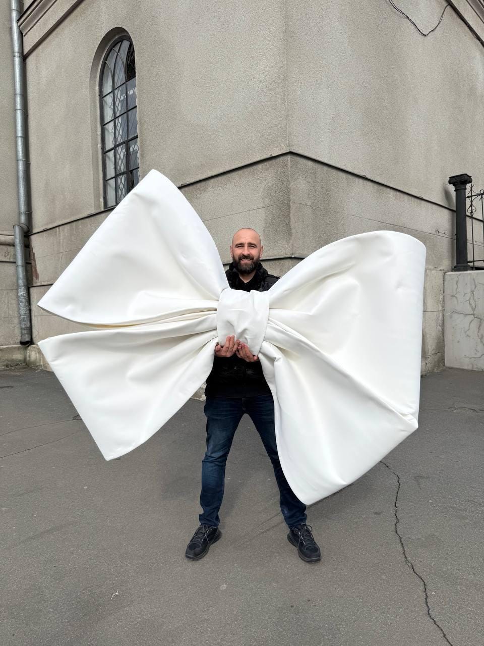 May include: A large white bow made of fabric, held by a person in front of a building.