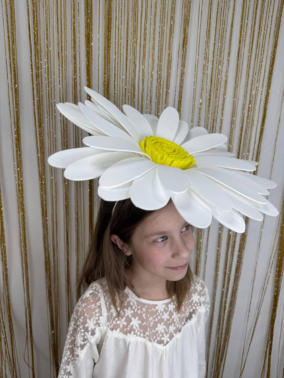 May include: A large, white daisy flower crown with a yellow center. The petals are layered and the crown is worn on the head. The background is a gold, glittery curtain.