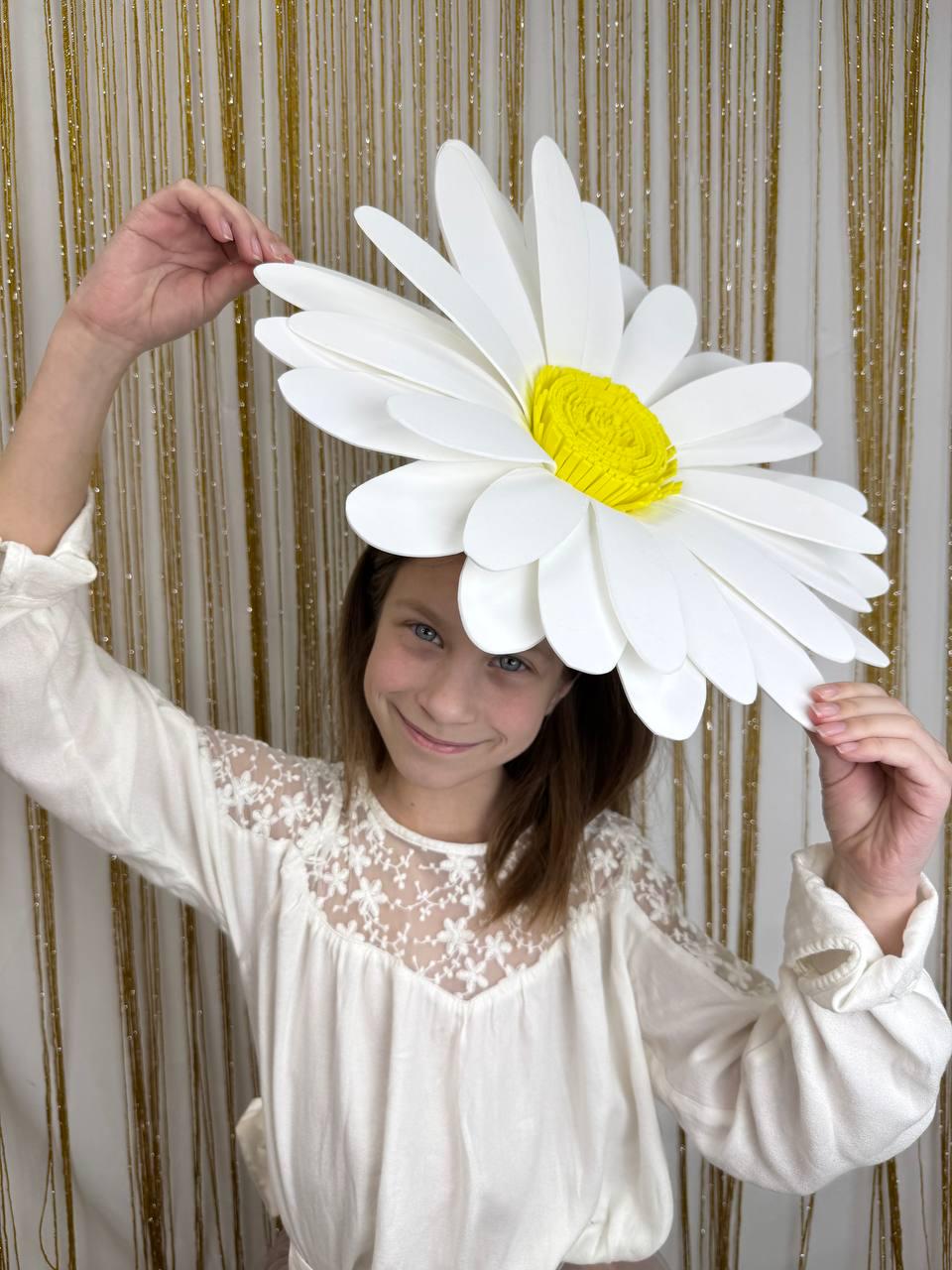 May include: A young person wearing a white shirt with lace detailing holds a large white paper daisy with a yellow center. The daisy is made of multiple layers of paper petals.