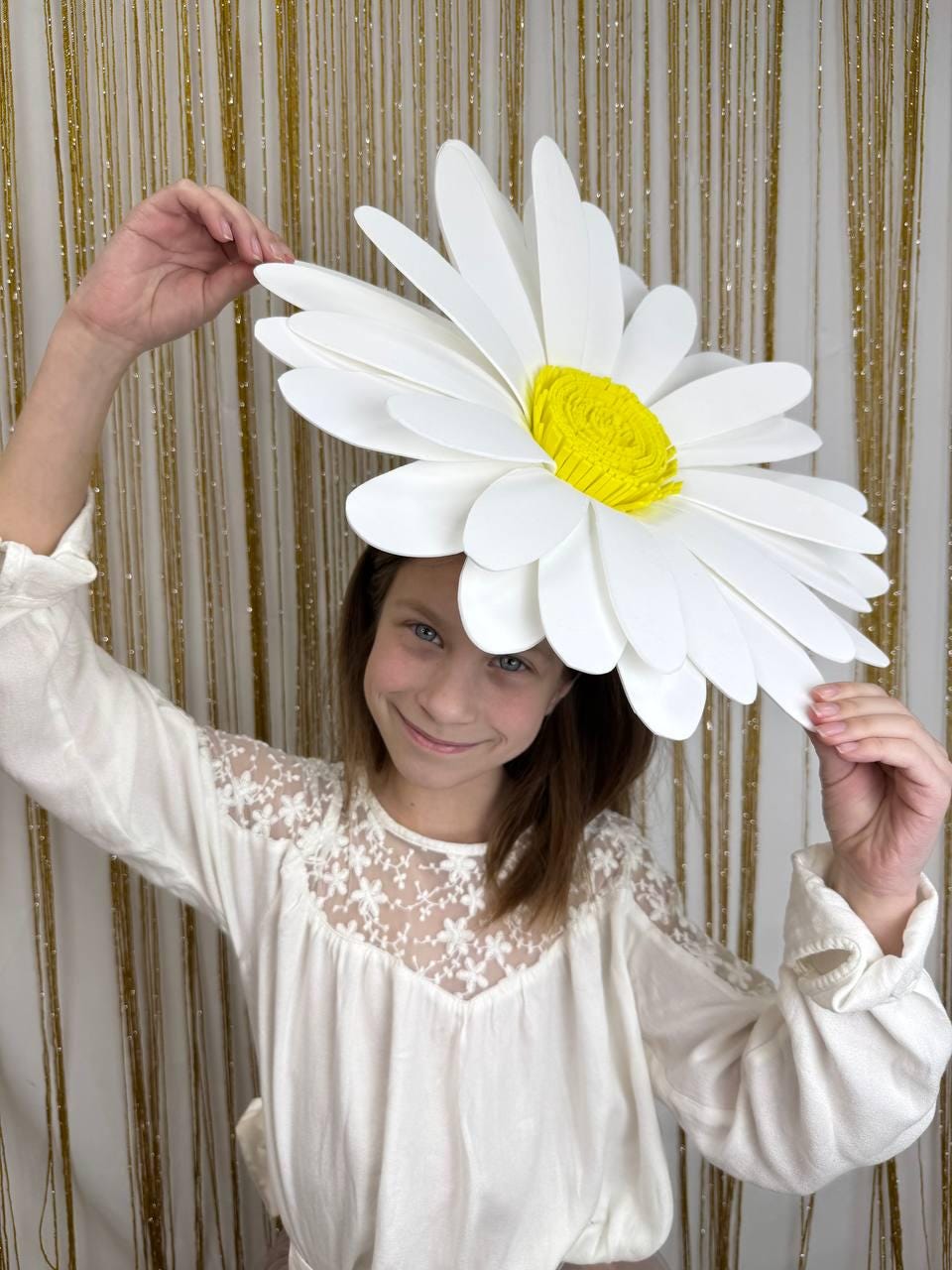 May include: A large, white daisy headpiece with a bright yellow center. The oversized flower is held up against a gold backdrop. The person is wearing a white blouse with lace detailing.