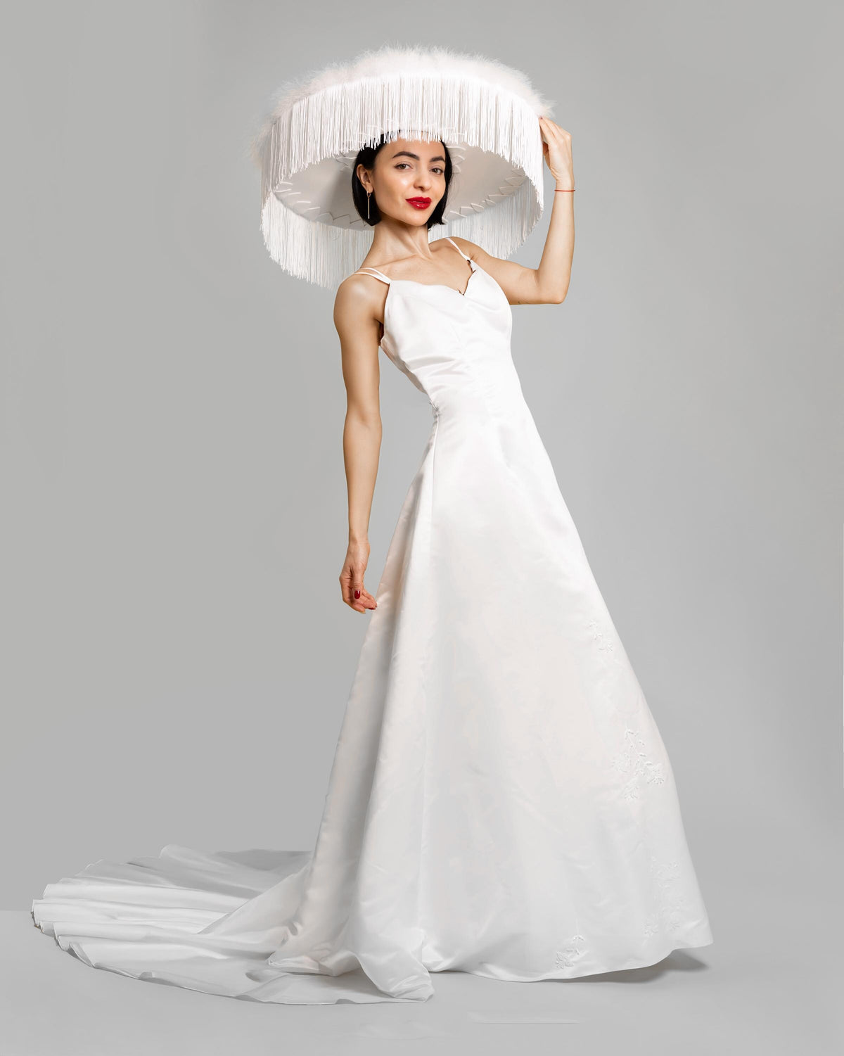 May include: A woman wearing a white wedding dress with a long train and a large white hat with a wide brim and feather trim.