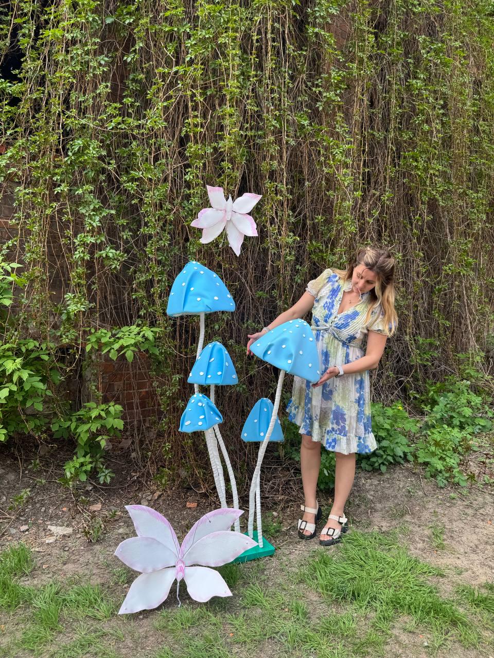 May include: A woman stands in front of a backdrop of green vines and holds a blue mushroom with white polka dots.  There are four other blue mushrooms with white polka dots in the background.  A white butterfly with pink accents is on the ground in front of the mushrooms.  A white flower with pink accents is in the background.