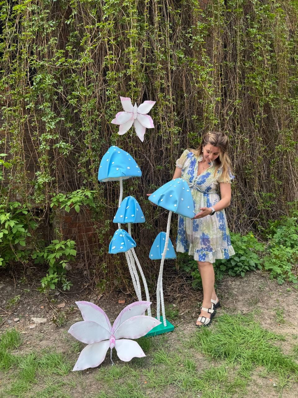 May include: A woman stands in front of a whimsical display of blue and white mushroom sculptures and butterflies. The mushrooms have white stems and blue tops with white polka dots. The butterflies are white with pink accents.